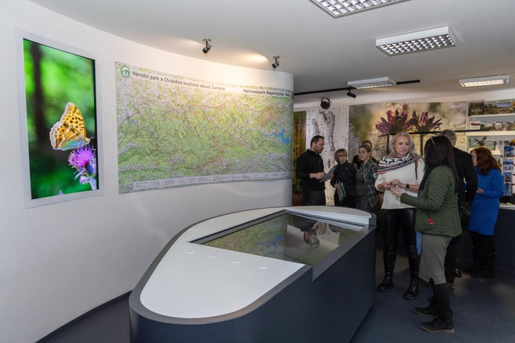 otevreni IS Kvilda po rekonstrukci, hosté slavnostní vernisáže debatují a na stěně za nimi je velkoformátová mapa území Šumavy
