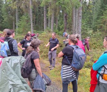 foto ze seminare Pojdme na to od lesa, účastníci naslouchají výkladu lektora, jsou na kraji lesa, v pozadí jsou stromy