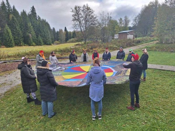 Účastníci semináře stojí v kruhu na louce u Lesní dílny a drží mezi sebou okraje padáku, na hlavách mají čepičky představující některá zvířata